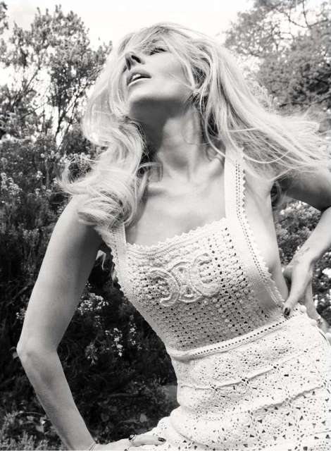 Monochrome portrait of a blonde adult woman with long wavy hair, wearing a crocheted dress, posing outdoors in a garden setting.