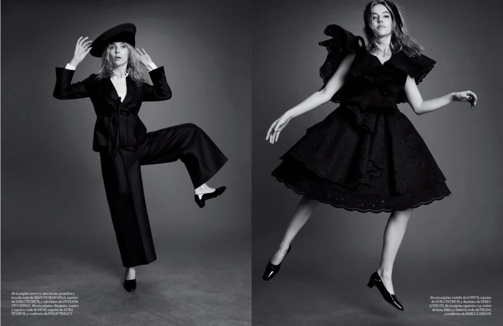Two women in black dresses and hats pose against a grey backdrop in a monochrome fashion photograph for a magazine spread.