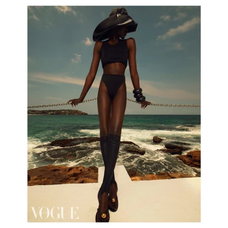 Model in black two-piece swimwear, wide-brim hat, and tall boots standing by the ocean by Emma Summerton