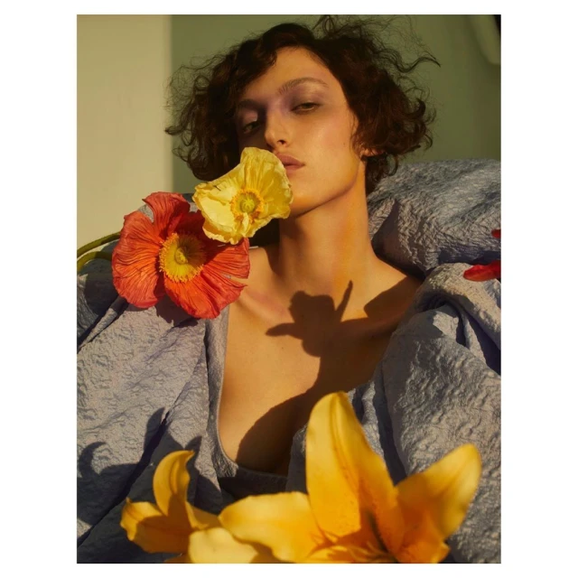 Portrait of a woman with short brown hair and light skin, wearing a periwinkle-blue textured dress, posing with orange and yellow flowers.