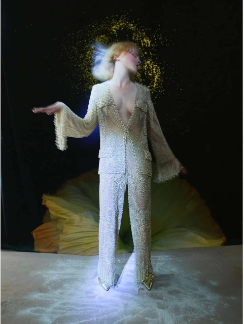 Woman in embellished pantsuit poses against a dark backdrop with a yellow spotlight effect, creating a high-fashion, studio-lit portrait.