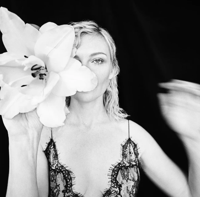 Black and white portrait of a woman with blonde hair, wearing a black lace top, posing with a white flower covering part of her face.