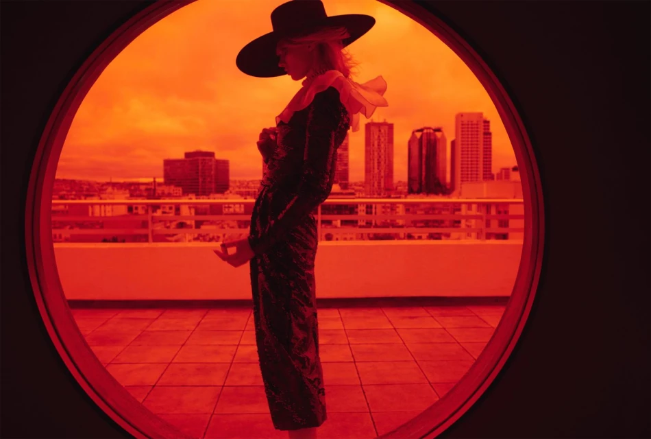 Woman in black hat and sequined dress stands on balcony framed by red circle, cityscape backdrop, elegant fashion photography, beauty shot.