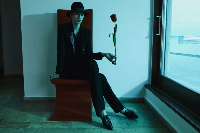 Man in black suit and fedora seated on a red chair holding a red tulip by Emma Summerton