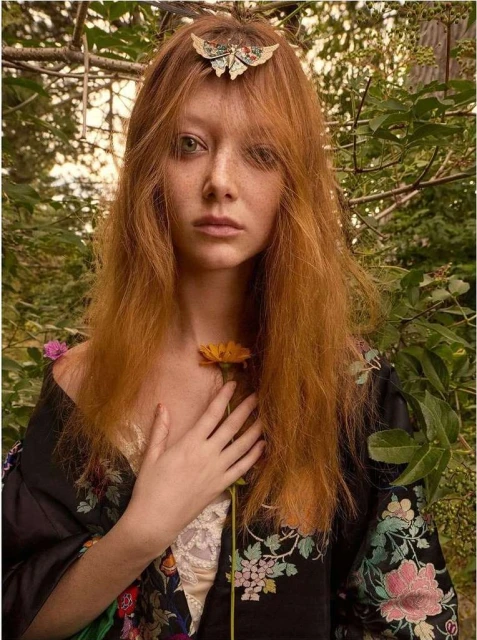 Portrait of a red-haired adult female wearing a floral robe, standing outdoors and holding a yellow flower, gazing calmly at the camera.