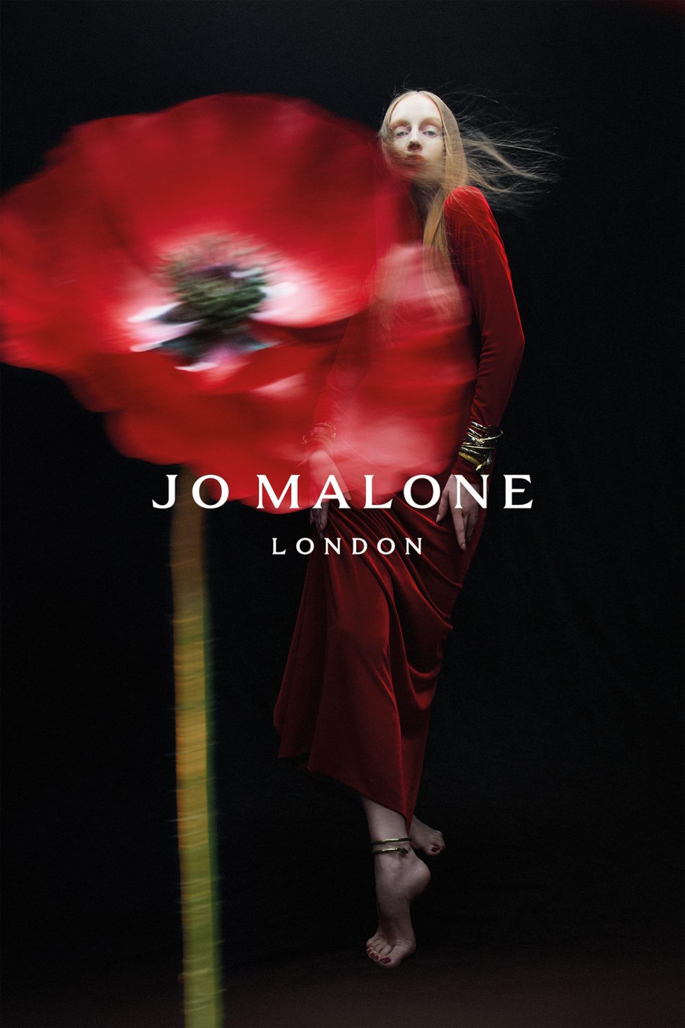 Jo Malone London campaign featuring an adult female in a red dress and bracelets, beside a blurred red flower against a black background.