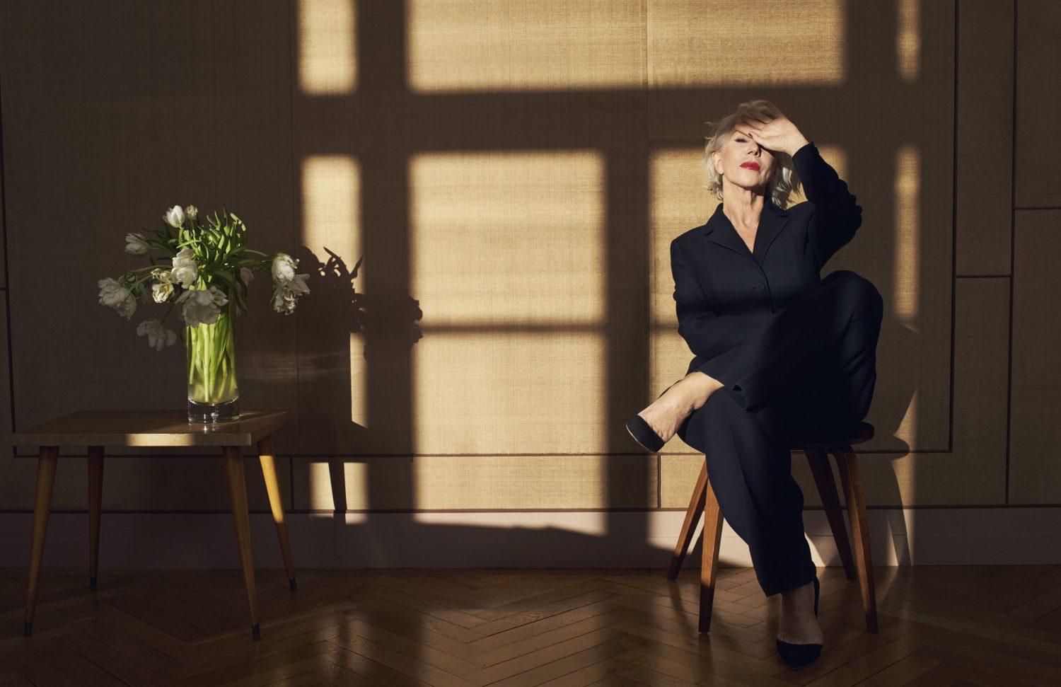 Mature woman in dark suit poses in sunlight near flowers, seated on a stool indoors with wood floors and neutral backdrop.