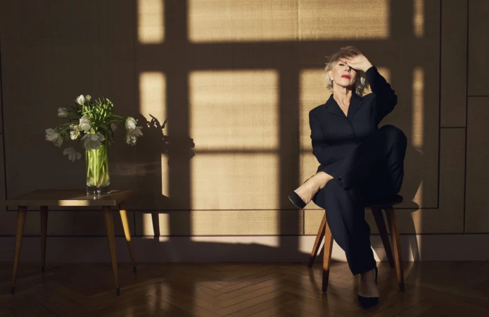 Mature woman in dark suit poses in sunlight near flowers, seated on a stool indoors with wood floors and neutral backdrop.