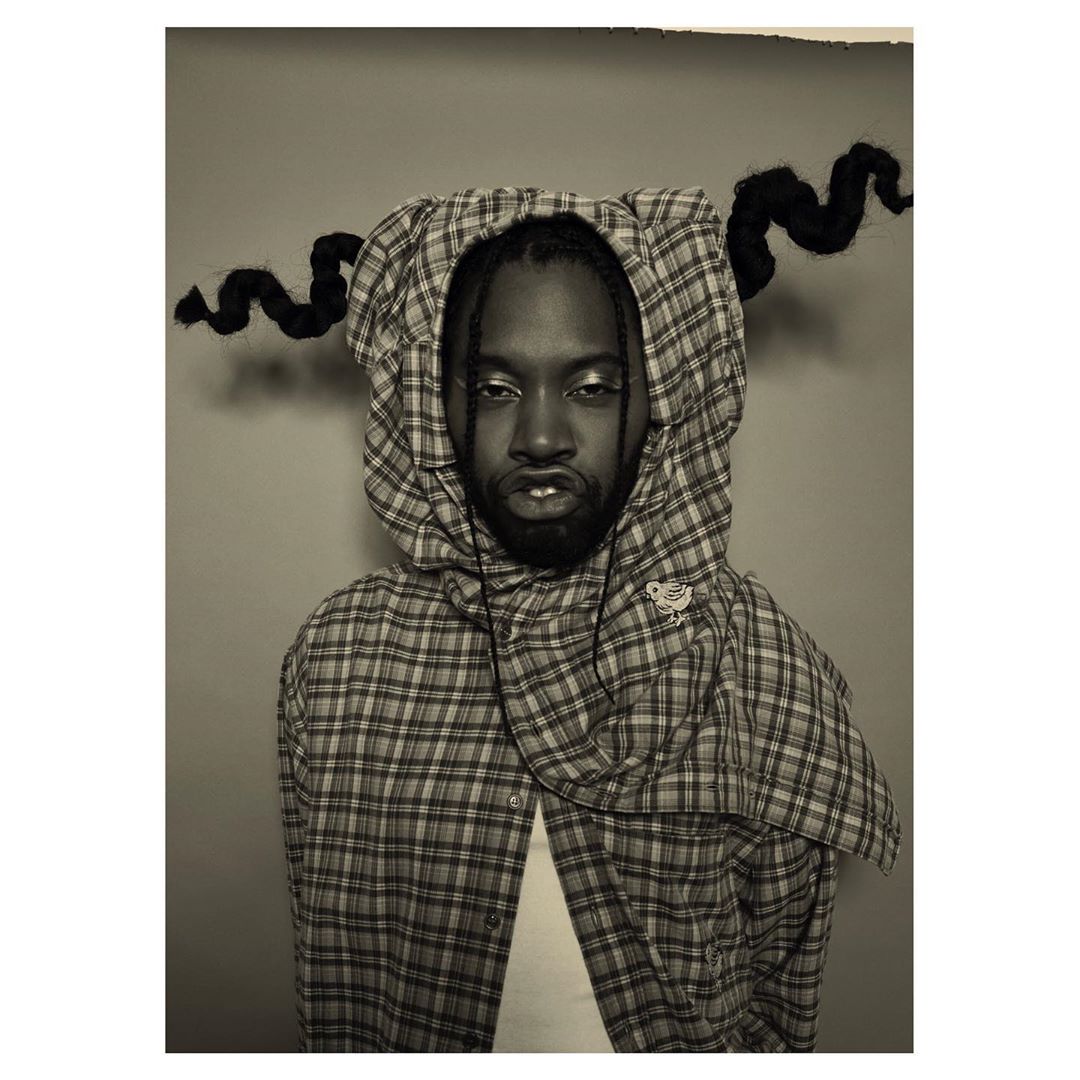 Portrait of a Black man wearing a plaid hooded shirt in a studio setting, featuring creative hair styling and makeup, captured in a medium shot.