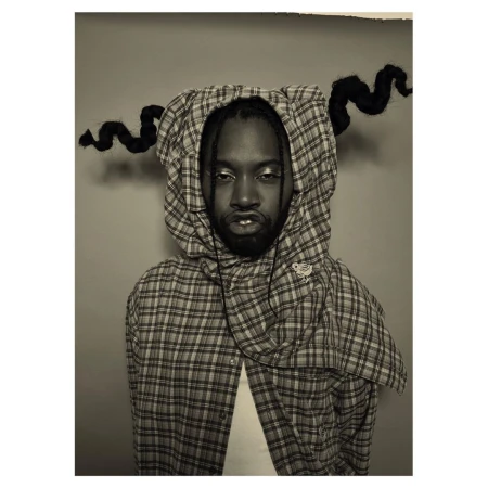 Portrait of a Black man wearing a plaid hooded shirt in a studio setting, featuring creative hair styling and makeup, captured in a medium shot.