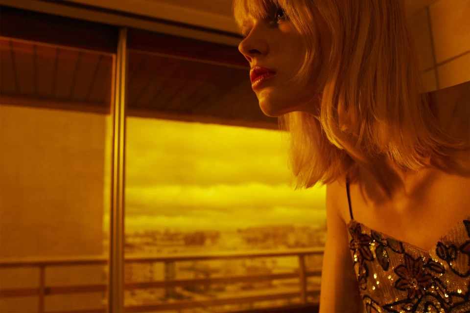 Woman with blonde hair and red lipstick posing indoors near a window with a city view in a yellow, warm-toned image.