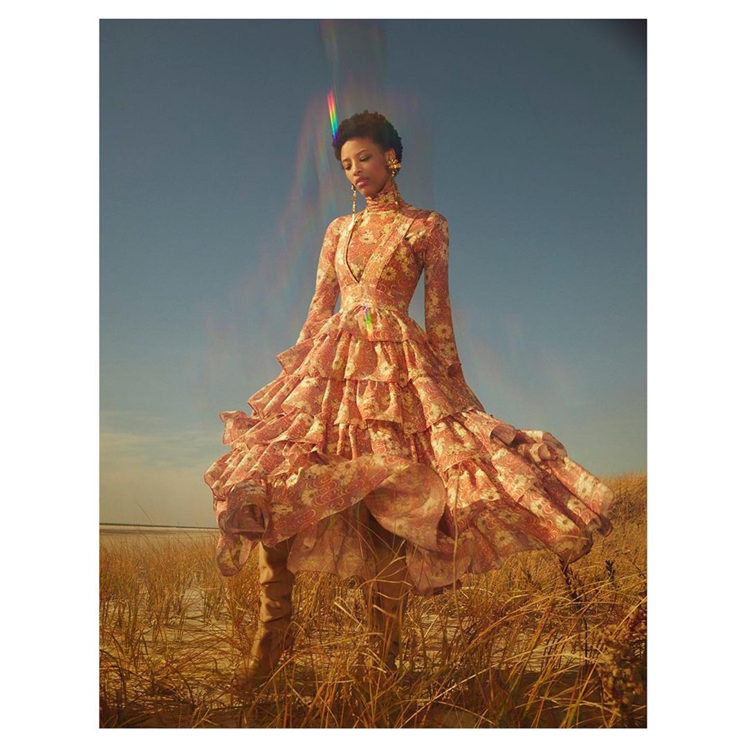 Young woman poses outdoors in a long-sleeved, floral-patterned, ruffled dress and tan boots against a blue sky with dry grass and water.
