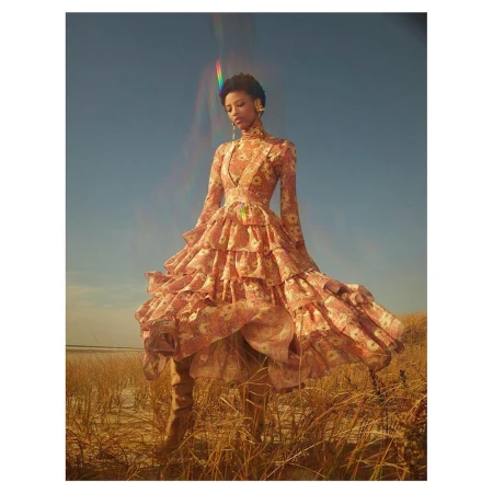 Young woman poses outdoors in a long-sleeved, floral-patterned, ruffled dress and tan boots against a blue sky with dry grass and water.