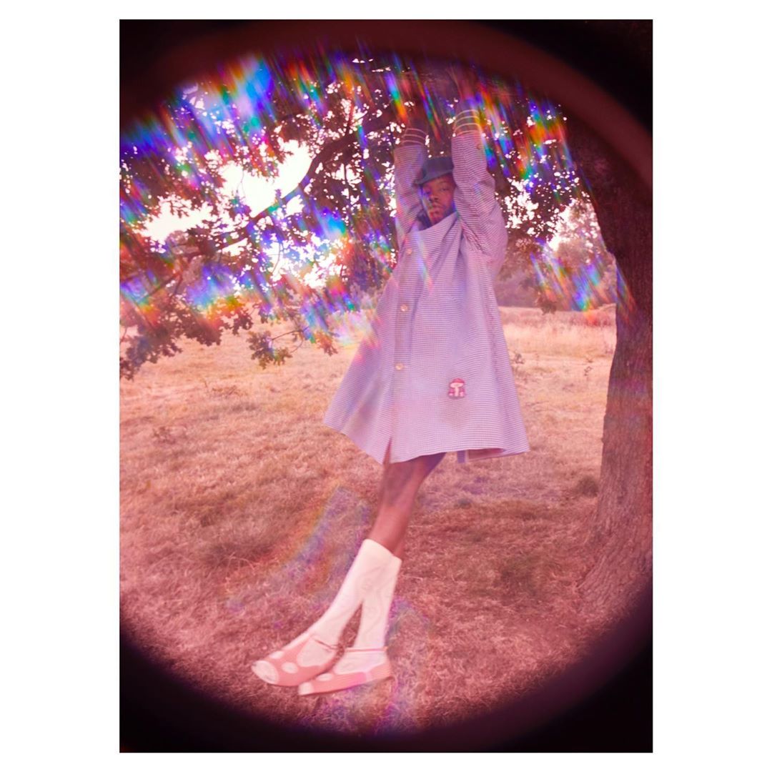 A person hangs from a tree branch wearing a light purple overcoat, white socks, and light pink sandals in a pastel-colored outdoor field.