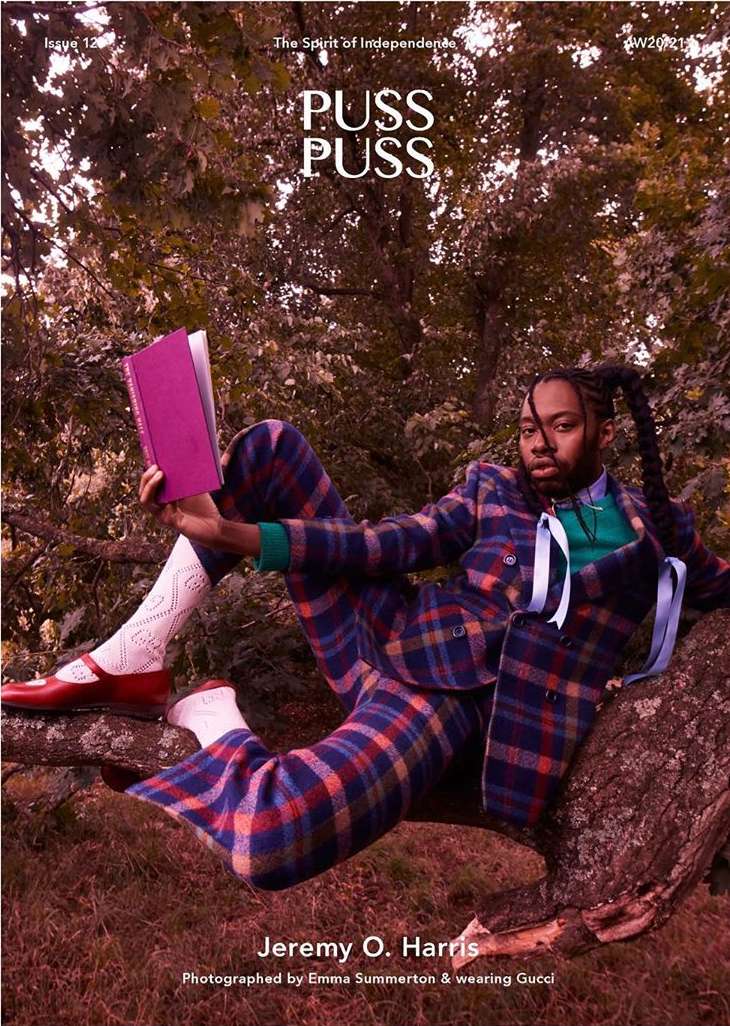 Man in plaid suit reads a book sitting on a tree branch outdoors. He wears red shoes and textured socks in natural light.