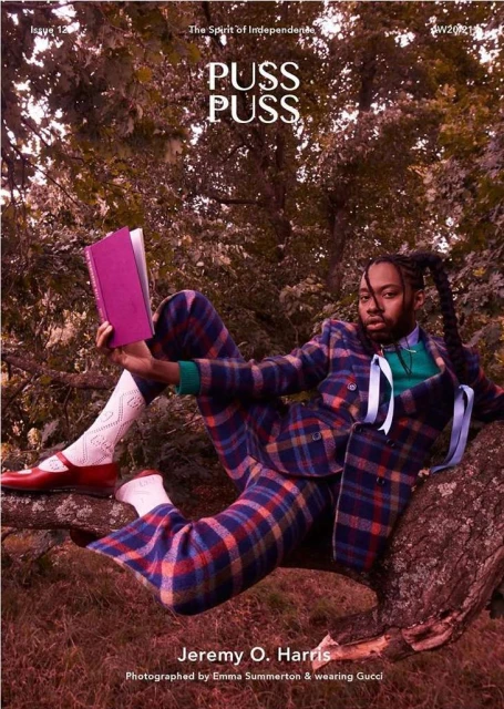 Man in plaid suit reads a book sitting on a tree branch outdoors. He wears red shoes and textured socks in natural light.