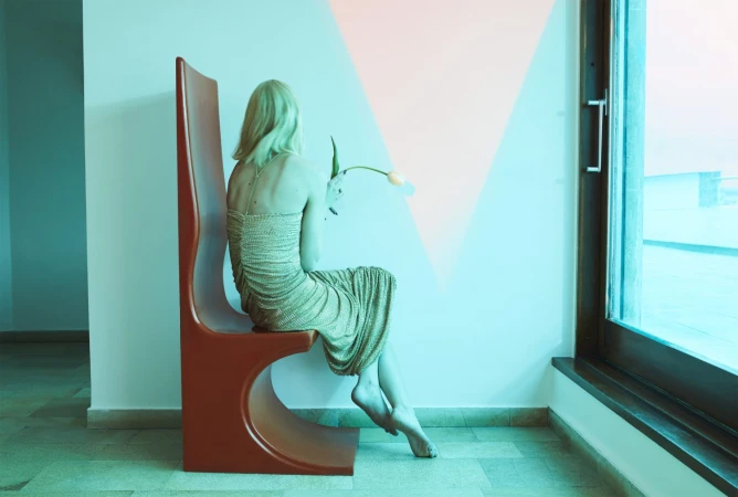 Blonde woman in a textured gold dress seated on a tall brown chair holding a single tulip near a large window by Emma Summerton
