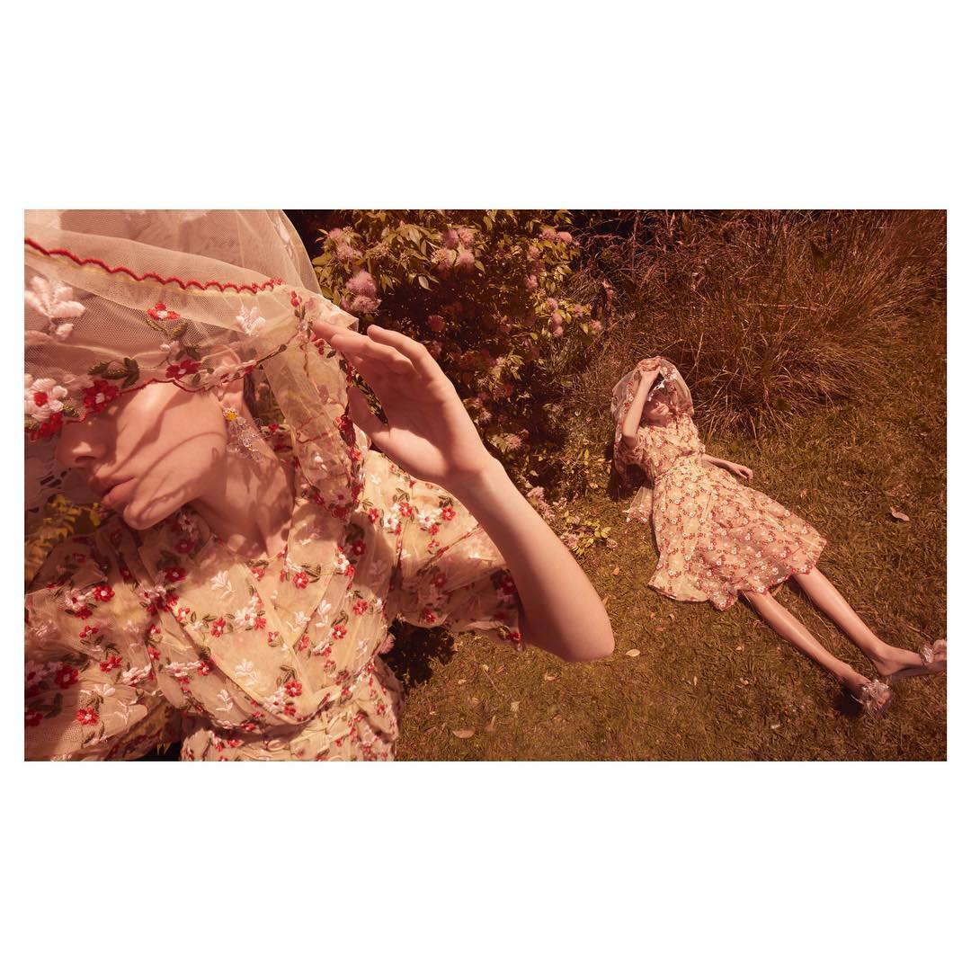 Two women in floral dresses, one lying on grass and the other partially visible, wearing a veil, under soft light and shadow.