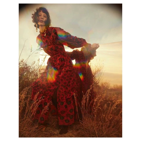 A woman in a flowing red dress stands in a field of tall grass under a cloudy sky with a rainbow light flare in the image.
