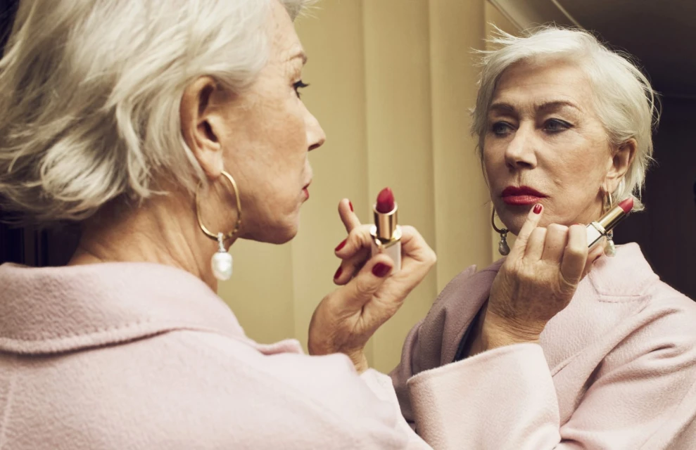Mature woman with short gray hair applies red lipstick in the mirror, wearing gold earrings and a pink coat in an indoor setting.