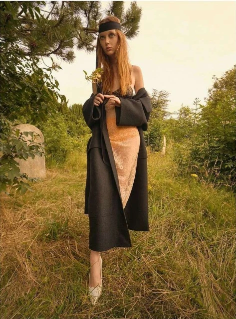 A long-haired redheaded woman with a black headband stands in a grassy field wearing a black coat, a tan dress, and heels holding a sprig.