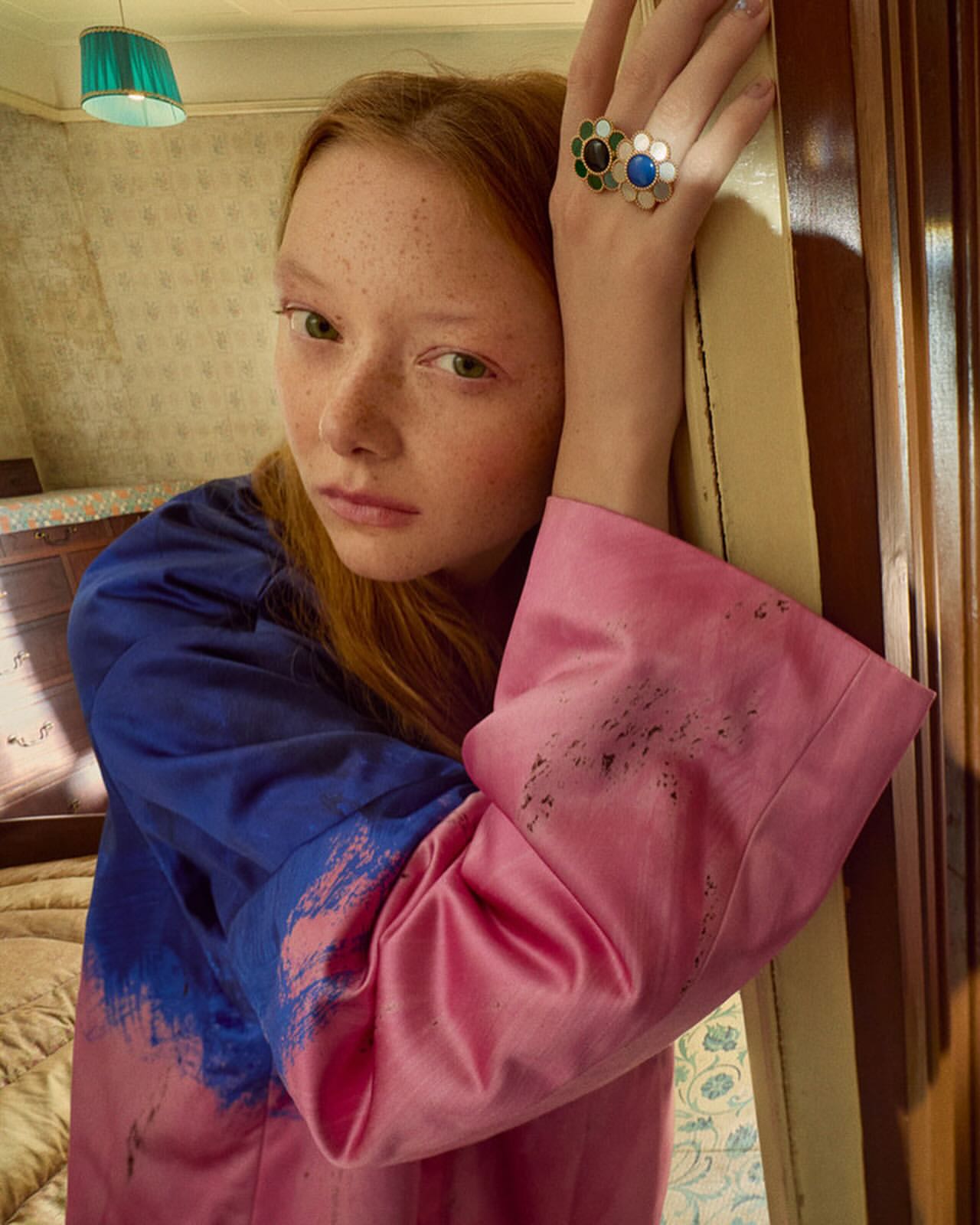 Close-up portrait of a red-haired woman leaning against a wooden doorframe wearing a blue and pink top and statement rings
