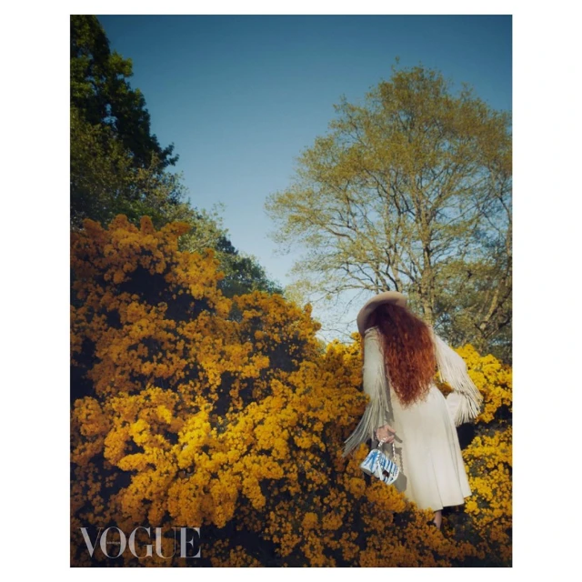 Fashion editorial shot of a red-haired woman in a coat with fringe, standing amidst golden gorse bushes under a clear blue sky, Vogue logo.