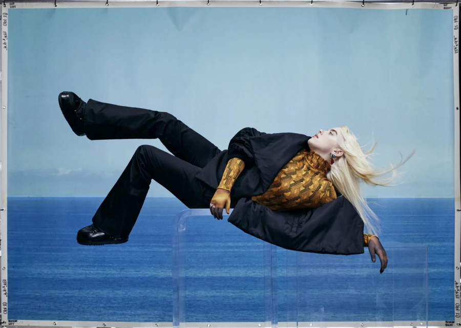 Woman lies on a clear platform in black suit with patterned blouse, set against a blue background for a fashion editorial.