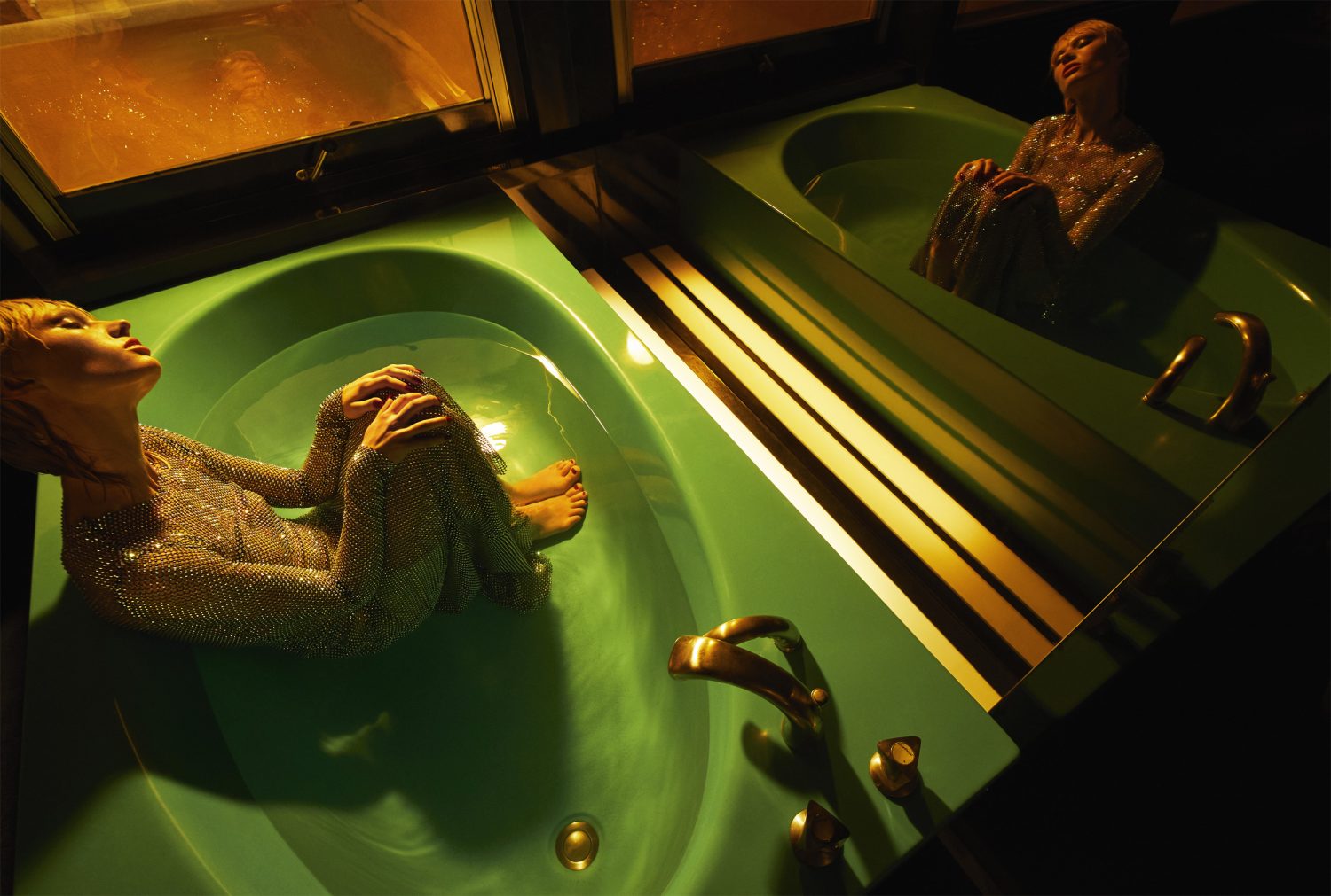 Two adult women recline in separate green bathtubs in a dimly lit indoor space with gold faucet fixtures and window light.