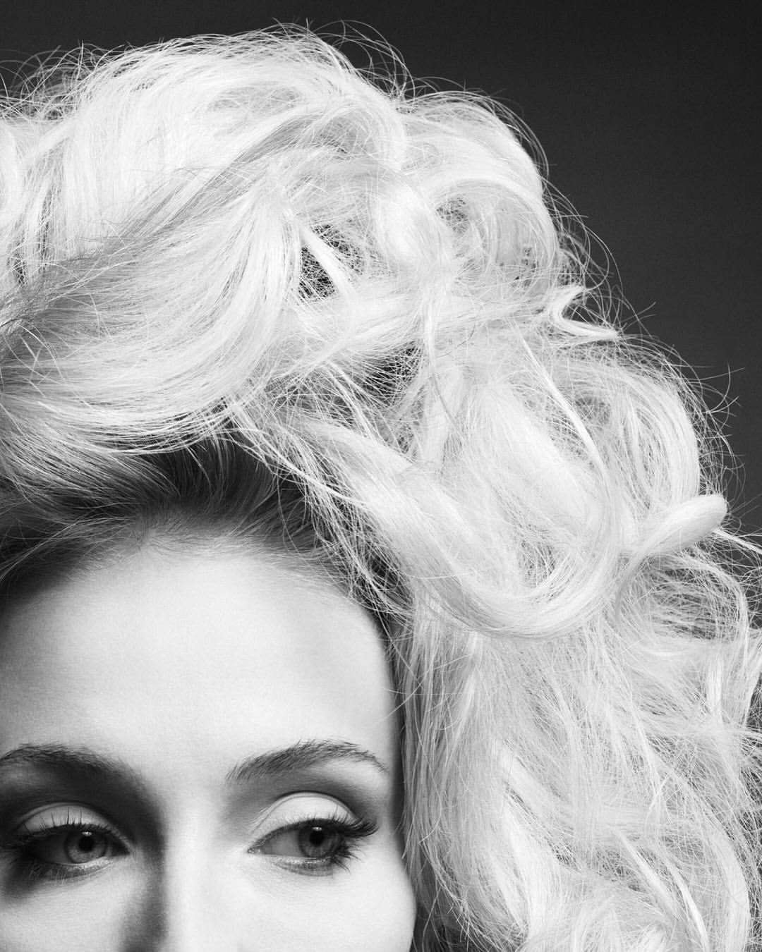 Close-up black and white shot of a woman with blond wavy hair and smokey eye makeup, set against a dark backdrop in a photo studio.