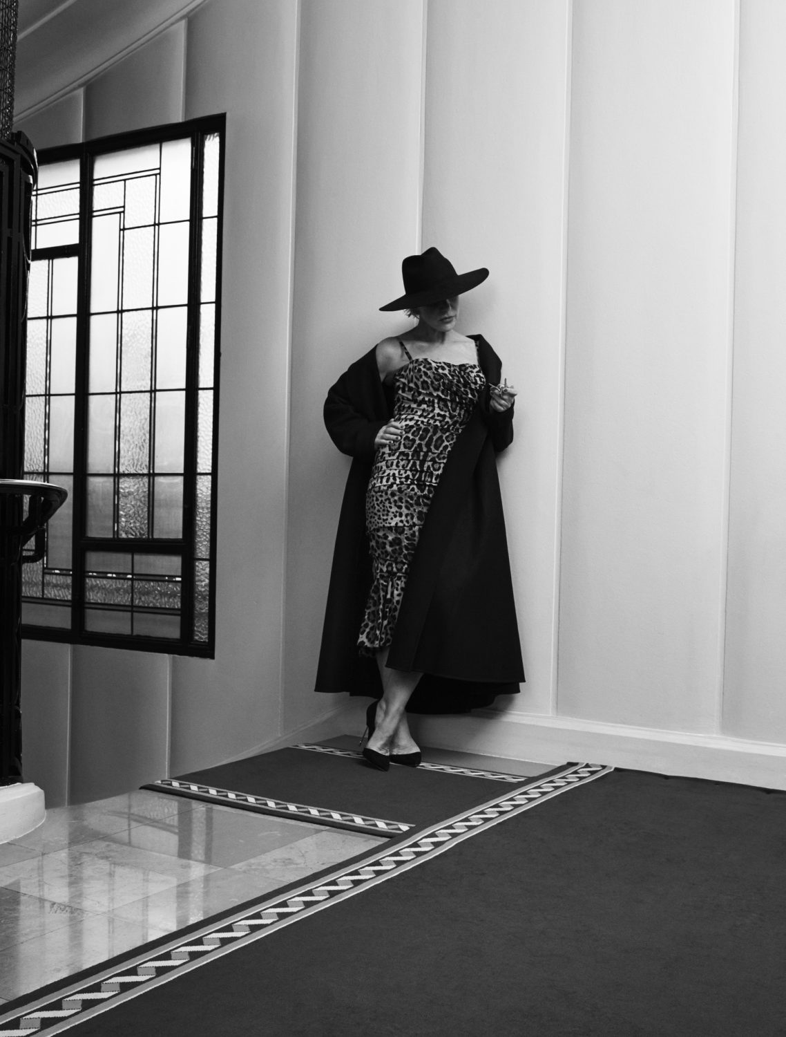 A woman in a leopard-print dress and black coat and hat poses in a corner with a window and patterned floor in a black-and-white photograph.