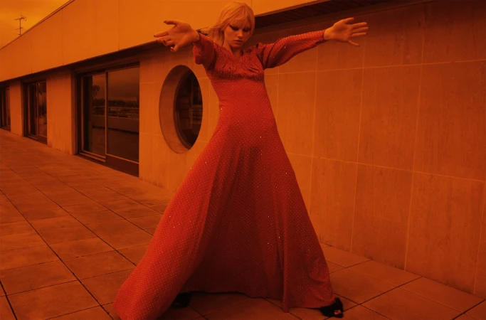 Model in long red evening dress with arms extended standing on a tiled terrace under warm orange light
