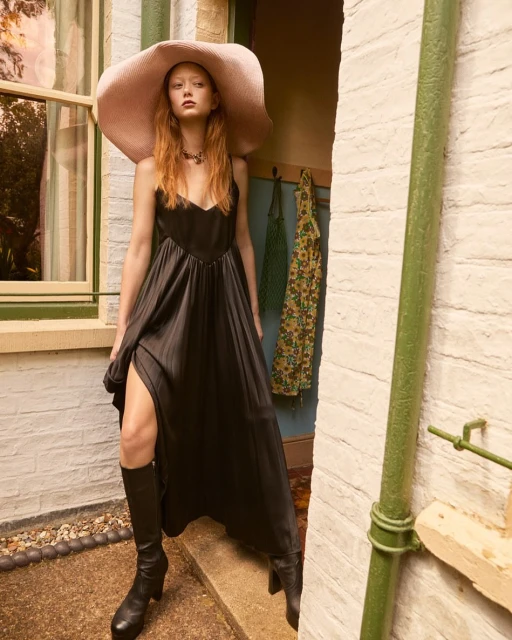 Elegant redhead woman in a black dress and oversized hat poses in a doorway with textured walls and cobblestone ground.
