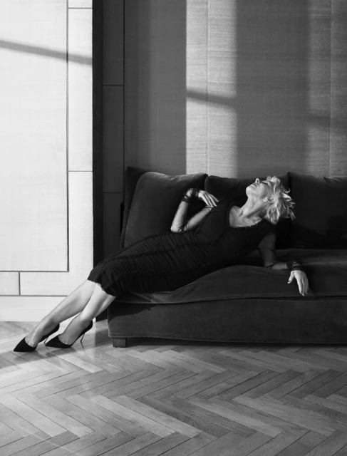 Elegant woman in black dress and heels lounges on a couch in a brightly lit room with herringbone wood floors and modern decor.