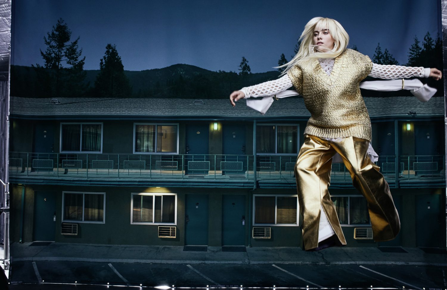 A blonde woman models in metallic gold clothing against a staged backdrop of a motel at night in a studio fashion shoot.