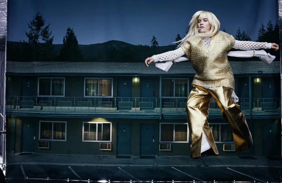 A blonde woman models in metallic gold clothing against a staged backdrop of a motel at night in a studio fashion shoot.
