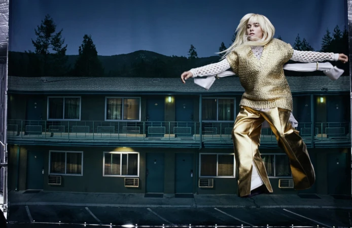 A blonde woman models in metallic gold clothing against a staged backdrop of a motel at night in a studio fashion shoot.