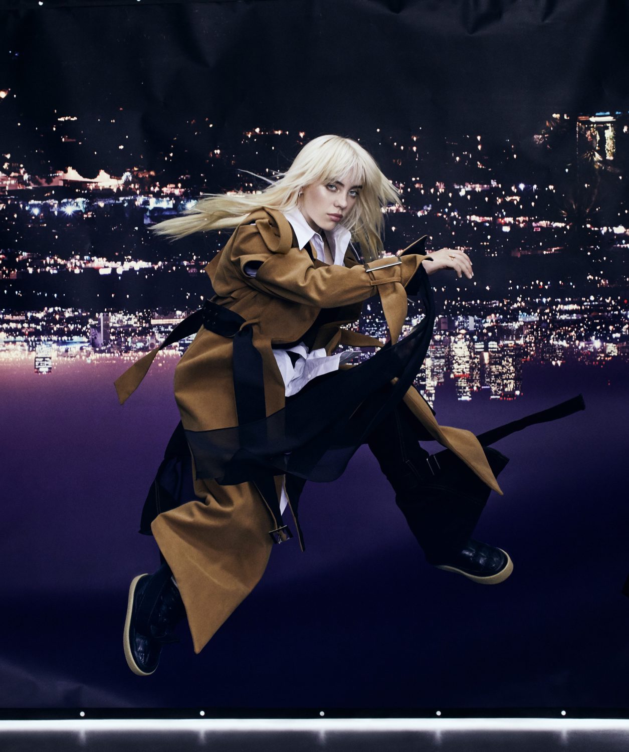 Blond female adult in motion, wearing a tan trench coat, black pants, and sneakers, posed in front of a city lights backdrop.