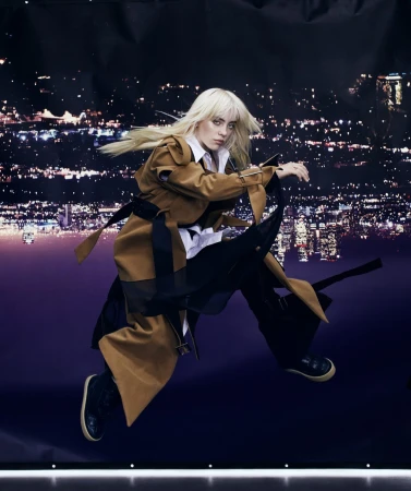 Blond female adult in motion, wearing a tan trench coat, black pants, and sneakers, posed in front of a city lights backdrop.