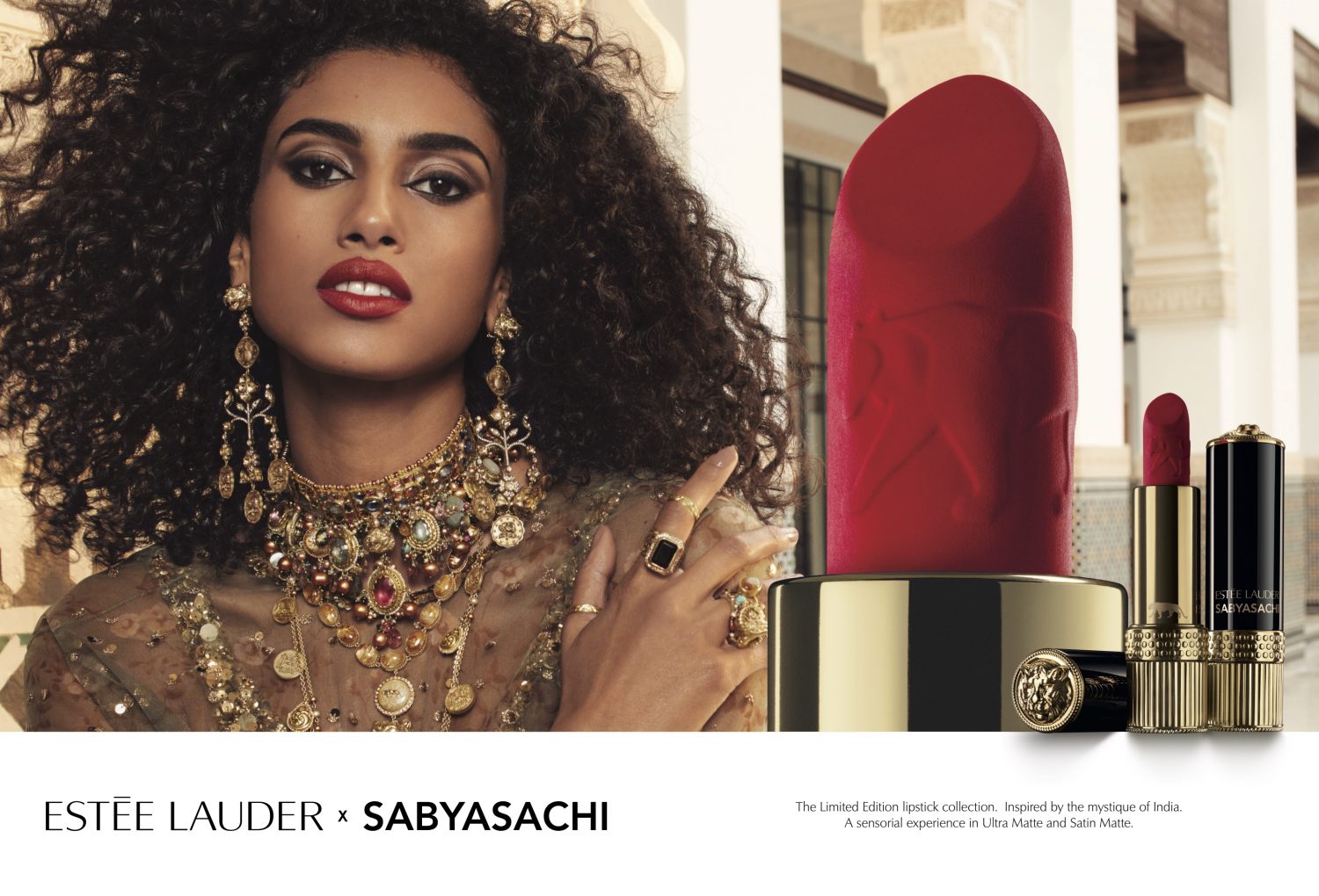 Elegant model with curly hair wearing jewelry and a red lipstick with Estée Lauder x Sabyasachi beauty products in a studio setting.