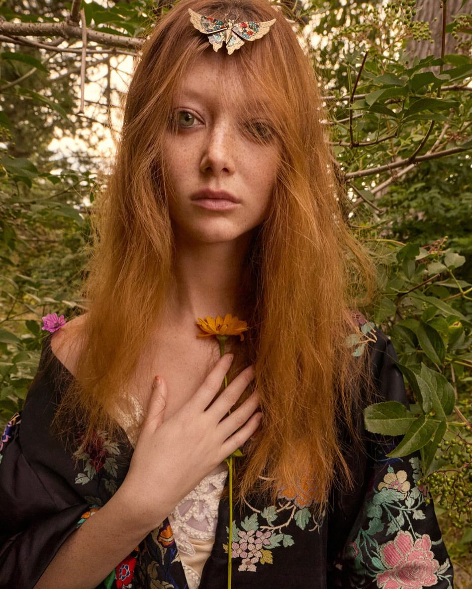 Portrait of a red-haired woman in an outdoor setting, wearing a floral kimono-style robe and holding a yellow flower in her hand.