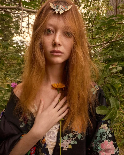 Portrait of a red-haired woman in an outdoor setting, wearing a floral kimono-style robe and holding a yellow flower in her hand.