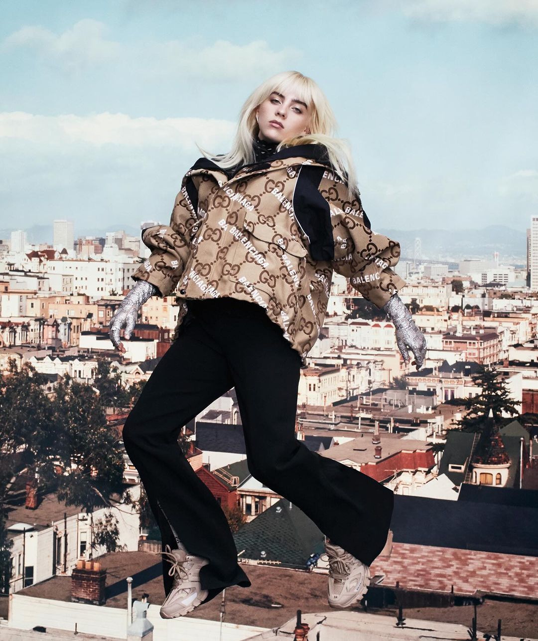 Blonde-haired woman in jacket and black pants strikes a pose in an urban setting with city buildings and a cloudy sky background.