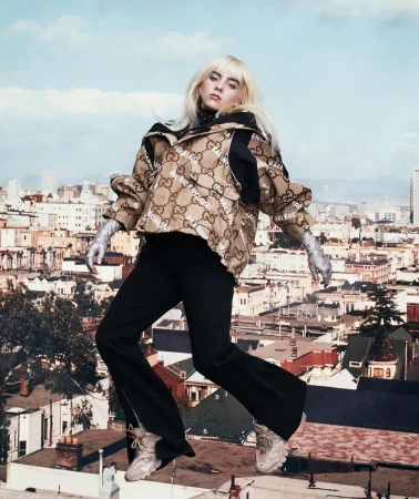 Blonde-haired woman in jacket and black pants strikes a pose in an urban setting with city buildings and a cloudy sky background.