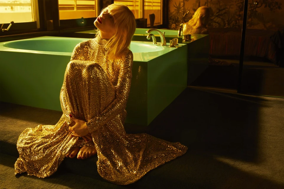 Adult female in long gold sequin gown sitting barefoot on the floor of a warm-toned interior with a mint-green tub and muted floral wallpaper.