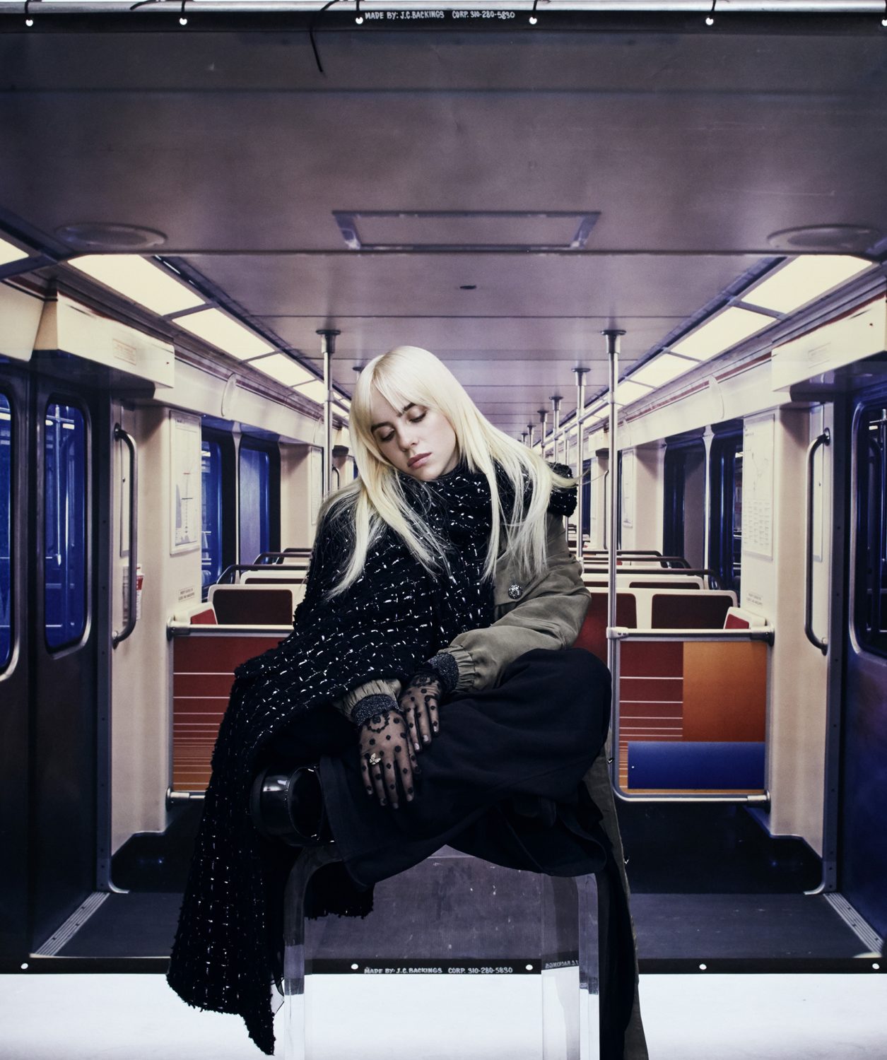 Blonde woman in a black and white scarf sits inside a subway car with empty seats, wearing a jacket and black pants.