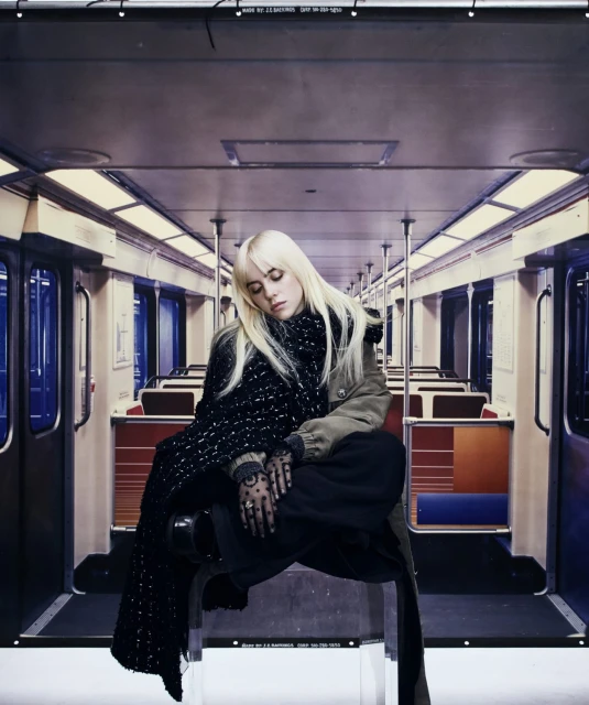 Blonde woman in a black and white scarf sits inside a subway car with empty seats, wearing a jacket and black pants.