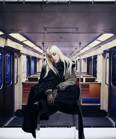 Blonde woman in a black and white scarf sits inside a subway car with empty seats, wearing a jacket and black pants.