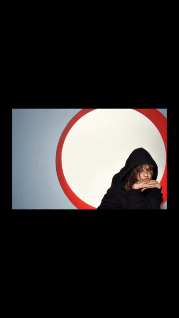 Woman with dark hair wearing a black hooded sweatshirt poses in front of a white circle with a red ring, against a grey background.