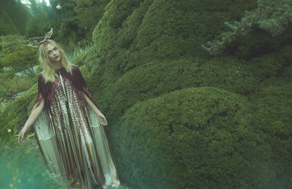 Woman in flowing gown with fringed cape and leaf headpiece poses in a lush green garden with rounded bushes and various plant life.